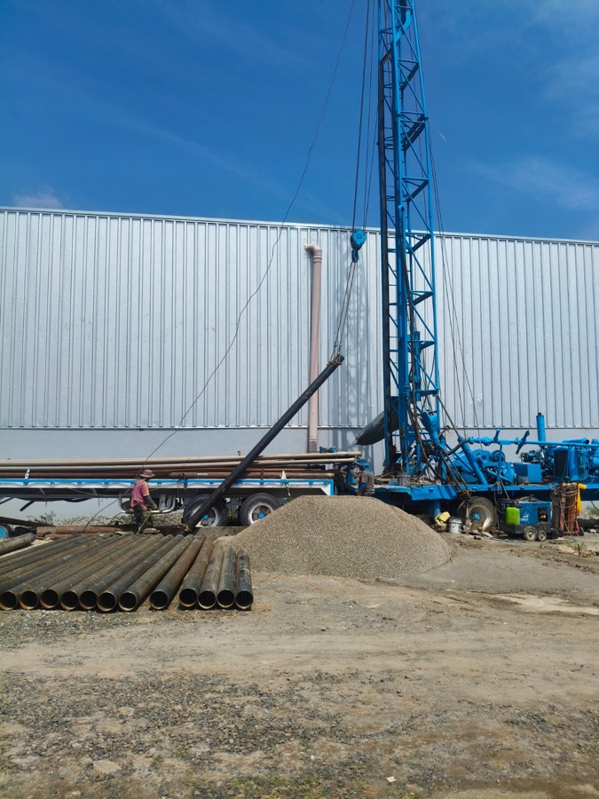 Equipo de perforación en operación junto a una nave industrial, con tubería de ademe y grava en el sitio